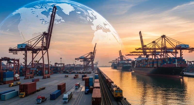 夕焼けの港に停泊する巨大なコンテナ船と、クレーン、貨物列車、トラックが並ぶ、世界規模の物流を象徴するイメージ画像。